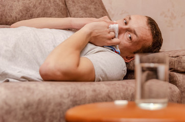 man sitting on the couch. He has a runny nose, so he blows her nose and wipes away tissue paper. place the medicine and a glass of water on the table next to it. The concept of the disease