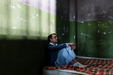 Depressed young man sitting on a mattress in a dark prison cell during custody