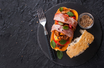 Stuffed pumpkin Butternut meat and vegetables wrapped in bacon with a slice of ciabatta on a black slate background. Copy space. Top view