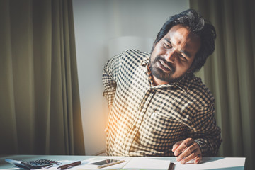 office syndrome concept; businessman sitting at office desk,  suffering from backache after working,man touching lower back with pained expression.