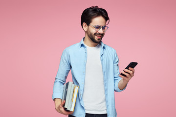 Young cheerful male student holding books and using a smart phone while standing isolated over pink background.