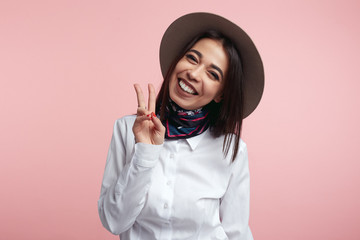 Pleased attractive woman shows v sign or peace gesture, wears white shirt and hat, isolated over rosy studio background. People, fashion, body language.