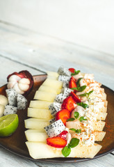 on a light wooden table lies a plate with a prepared meal of exotic fruits: honey melon, rambutan, dragon fruit, mangosteen with the addition of chilli peppers, strawberries and whipped cream