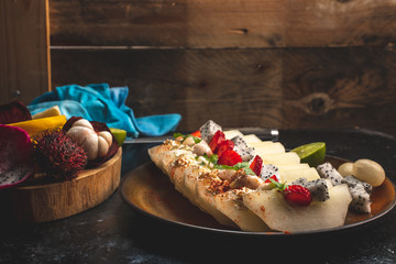 on a dark, stone table lies a plate with a prepared meal of exotic fruits: honey melon, rambutan, dragon fruit, mangosteen with the addition of chilli peppers, strawberries and whipped cream, sprinkle