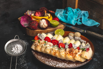 on a dark, stone table lies a plate with a prepared meal of exotic fruits: honey melon, rambutan, dragon fruit, mangosteen with the addition of chilli peppers, strawberries and whipped cream, sprinkle