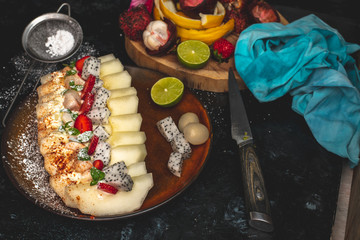 on a dark, stone table lies a plate with a prepared meal of exotic fruits: honey melon, rambutan, dragon fruit, mangosteen with the addition of chilli peppers, strawberries and whipped cream, sprinkle