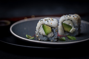 avocado and cream cheese inside out maki sushi wuth sesame seeds on dark plate with chopsticks and soy sauce