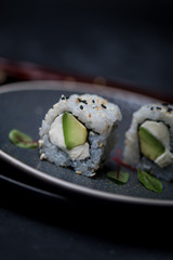 avocado and cream cheese inside out maki sushi wuth sesame seeds on dark plate with chopsticks and soy sauce
