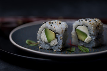 avocado and cream cheese inside out maki sushi wuth sesame seeds on dark plate with chopsticks and soy sauce