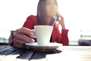 Young woman holding a hot cup of coffee while talking with her mobile phone sitting in the terrace of coffe shop.