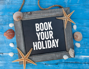 Chalkboard with maritime decoration on blue wooden background with Book your Holiday