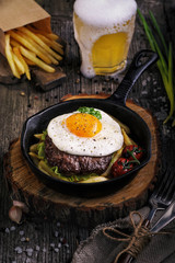 Fried minced beef steak with scrambled eggs, fried potatoes and tomatoes in a cast iron pan. Beer in a glass. Wooden table.
