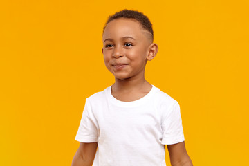 People, childhood, joy and happiness concept. Joyful cheerful Afro American boy of school age having fun, being in good mood, playing after he did his homework, posing isolated at yellow wall - obrazy, fototapety, plakaty