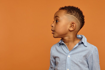 Waist up up image of handsome adorable dark skinned little child with stylish hairdo having emotional facial expression, standing isolated at blank orange wall with copyspace for your advertisement