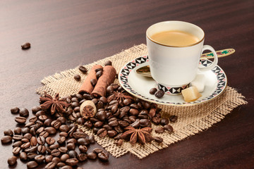 Coffee in porcelain cup on saucer with cinnamon, nutmeg and star anise on jute napkin. Vintage coffee set on wood table with scattered coffee beans.