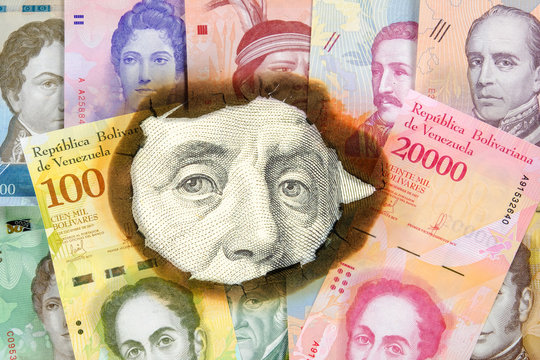 Concept of hyperinflation Venezuela. Burned Venezuelan money with the image of statesmen, through hole in which you can see an unburned one hundred dollar bill with Franklin. Close up.