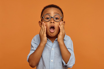 Horizontal image of funny surprised adorable black little boy in round glasses and shirt having...