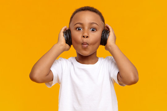 Picture of stylish cool Afro American schoolboy making funny facial expression, being excited with amazing track while listening to music using wireless black headphones, posing isolated in studio