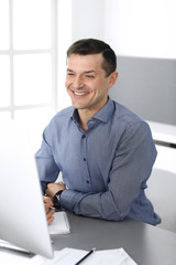 Naklejka premium Cheerful smiling businessman working with computer in modern office. Headshot of male entrepreneur or director of a company at the workplace. Business concept 