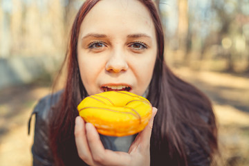 Young woman eating doughnut in woods Casual young brunette in jacket sitting on log in woods and eating delicious glazed doughnut