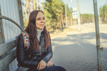 Young brunette sitting on bus stop Casual woman in black leather coat sitting on bus stop waiting for public transport looking away