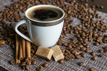 A Cup of coffee with foam stands on the table among the scattered coffee beans. Next to the Cup are pieces of sugar and cinnamon sticks.
