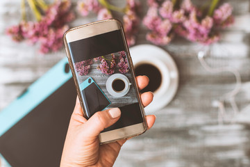 Blogger photographer workshop concept, hand holding a phone and taking photos of stylish flowers, a cup of coffee and a tablet computer on a gray wooden rustic background