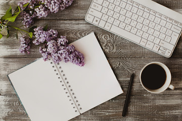 Top view computer keyboard, blank notebook and cup of coffee and lilac flowers on gray wooden table.