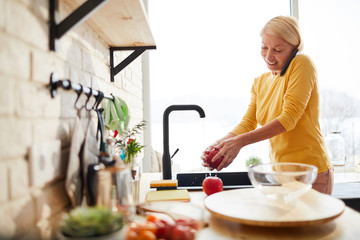 Cheerful attractive mature woman with blond hair standing at kitchen counter and washing fruit in sink while talking on mobile phone - Powered by Adobe