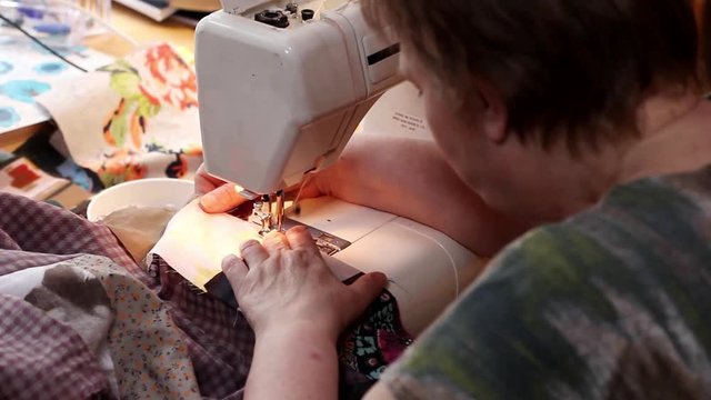 Mature woman sews patchwork quilt on sewing machine, focus on machine foot. Left-side view.