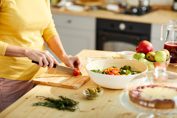Close-up of unrecognizable woman in yellow sweater standing at kitchen counter and cutting tomato while cooking Greek salad