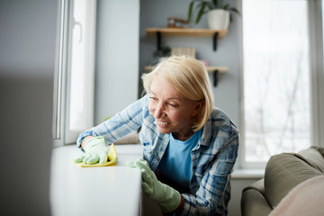Smiling satisfied mature woman with short blond hair wearing rubber gloves dusting window-sill with...