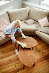 Fototapeta na wymiar Directly above view of blond-haired lady in rubber gloves sitting on table and cleaning wooden coffee table in living room, she keeping clean at home