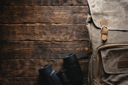 Travel or adventure flat lay background with copy space. A backpack and a binoculars on a brown wooden table.