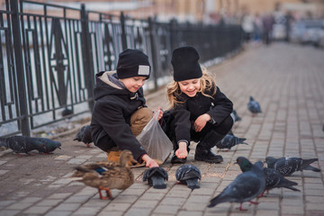 cute children, a brother and sister of 7-8 years old, in winter or early spring feed pigeons in the town square, wearing dark coats and hats
