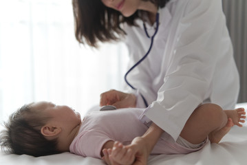Female doctor is listening heart pulse rate of cute newborn baby on the bed by using stethoscope in the room. Seen from side view of baby. Baby health care concept.