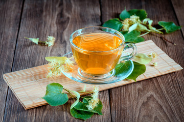 Cup of medicinal, lime tea, standing on a wooden table, surrounded by fragrant linden flowers, in the rays of sunlight.