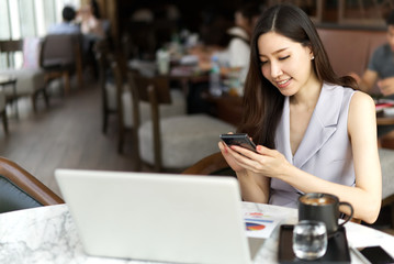 Smart working woman concept. Asian beautiful girl sitting in coffee shop and using mobile phone with smile to contact customer. Laptop and some drinks are put on the table.