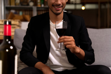 Luxury alcohol drinking. Front view of crop shot of man sitting on sofa holding glass of wine with smile before drinking. Bottle of wine put on table as foreground. Alcohol drinking and party concept.