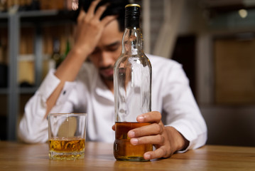 Drunk man sitting at the bar in pub and holding whisky bottle in left hand and put another hand on his head after he drank a lot of alcohol and feeling headache. Alcoholic concept.