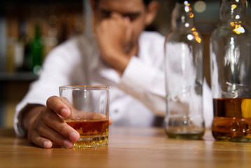 Drunk man sitting at the bar in pub and holding whisky glass in right hand and put another hand on his head after he drank a lot of alcohol and feeling headache. Alcoholic concept.