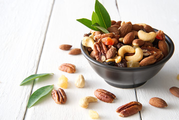 Mix nuts and dried fruits in the bowl put on the white painted wooden. Decorated with green leaf and some nuts on the wood. Seen from side view. Food and snack concept.