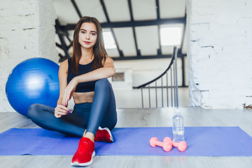 Pretty girl sitting on her mat with blue ball and dumbbells looking to the camera.
