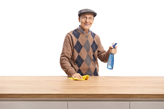 Elderly man with a cloth and cleaning spray standing behind a wooden counter and smiling at the camera