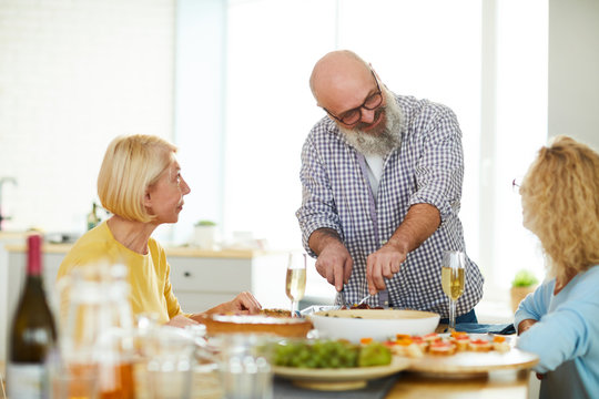 Content handsome mature bearded man in glasses standing at table and cutting meat for guests while talking to charming ladies at home