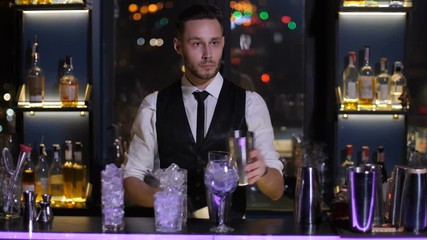 Handsome barman scooping ice cubes and putting in shaker during cocktail drink preparation in bar. Various glasses for alcoholic beverages full of ice in front of barkeeper at bar counter