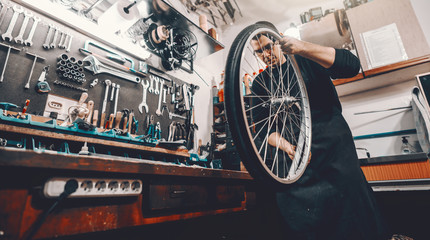 Serious handsome Caucasian man holding bicycle wheel in hands while standing in workshop.