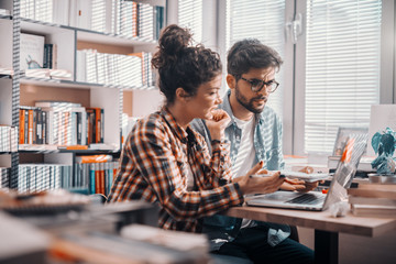 Obraz premium Two multicultural students studying for exams and using laptop while sitting in library. In background shelves with books.