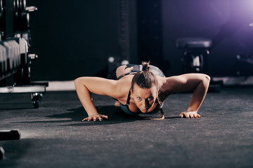 Fototapeta na wymiar Caucasian muscular woman with pigtail and in sportswear doing push ups on gym floor and looking at camera. Night workout concept.