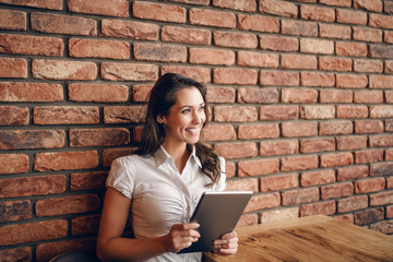 Smiling cute Caucasian brunette sitting in cafe and using tablet. In background brick wall.